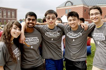 Five smiling students in matching grey shirts with arms around each other's shoulders on the Bardeen Quad.
