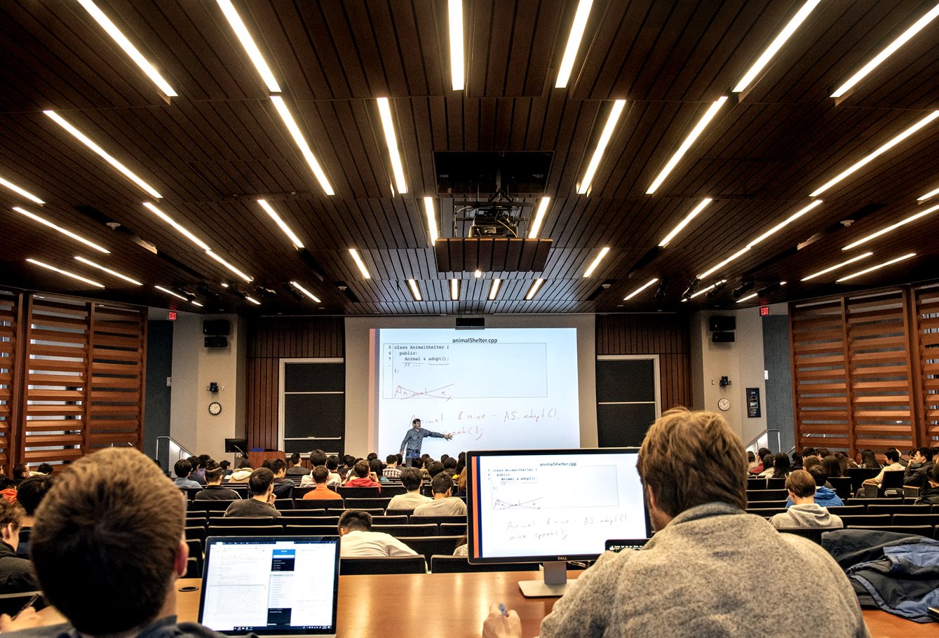 Large classroom with a professor at the front, students on laptops, and thin vertical fluorescent lights on the ceiling.