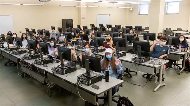 Classroom of students in front of computers, waving to camera