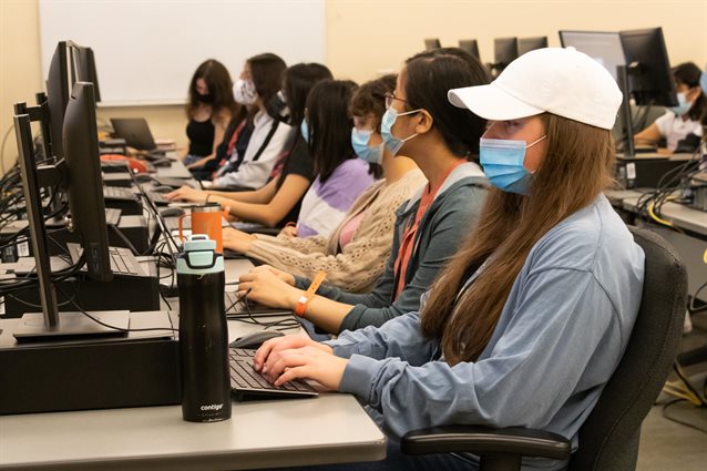 A row of students sitting at computers, viewed from the side. They are all facing forward.