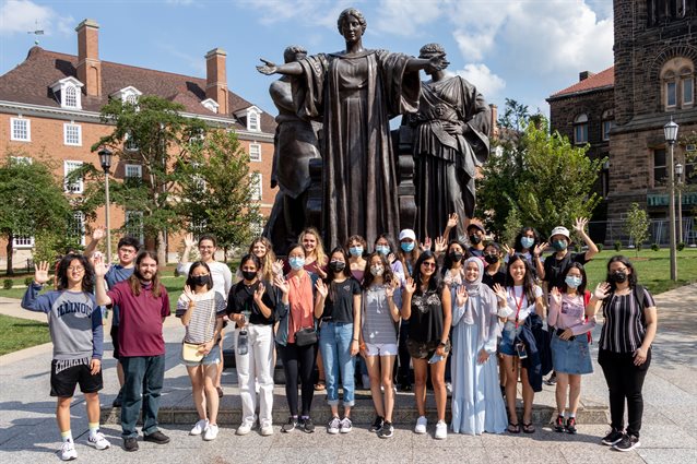 A group of students standing and waving in front of the Alma Mater statue.