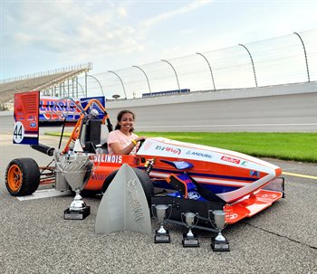 Illinois CS student and Illini Motorsports team member, Labdhi Jain, sits in the winning car designed by this team on a racetrack with trophies surrounding the car. The car is mostly orange and white with some blue.