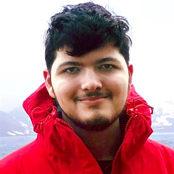 Headshot of recent alumnus Vivek Nair, outdoors in front of a body of water with a mountainous terrain in the background. Nair is wearing a winter coat.