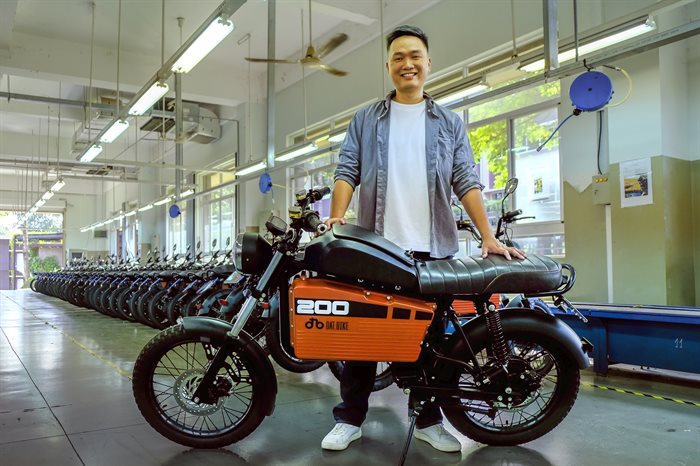 Illinois CS alumnus and current founder &amp;amp;amp; CEO of the company Dat Bike, Son Nguyen, stands in front of one of his the electric motorbikes his company produces. The picture is in a showroom with a line of the bikes, which have a black seat and an orange and black body.