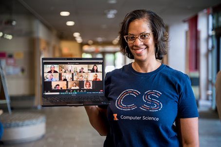 A woman smiles and holds a laptop displaying online students in the Siebel Center for Computer Science lobby.