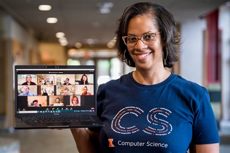 A woman smiles and holds a laptop displaying online students in the Siebel Center for Computer Science lobby.