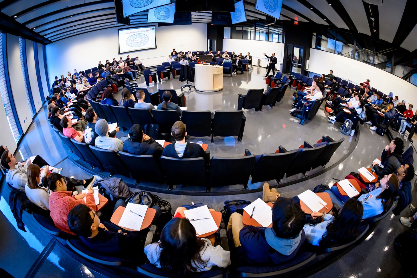 Students work and discuss design projects in a round classroom with numerous monitors hanging from the ceiling and a central podium designed to allow faculty and students to see each other more clearly.