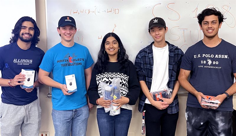 Five computer science students stand against a white board and hold small prizes.
