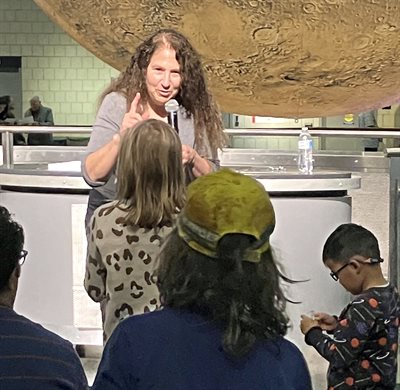 A woman stands with a microphone in a musem and talks to younger people.
